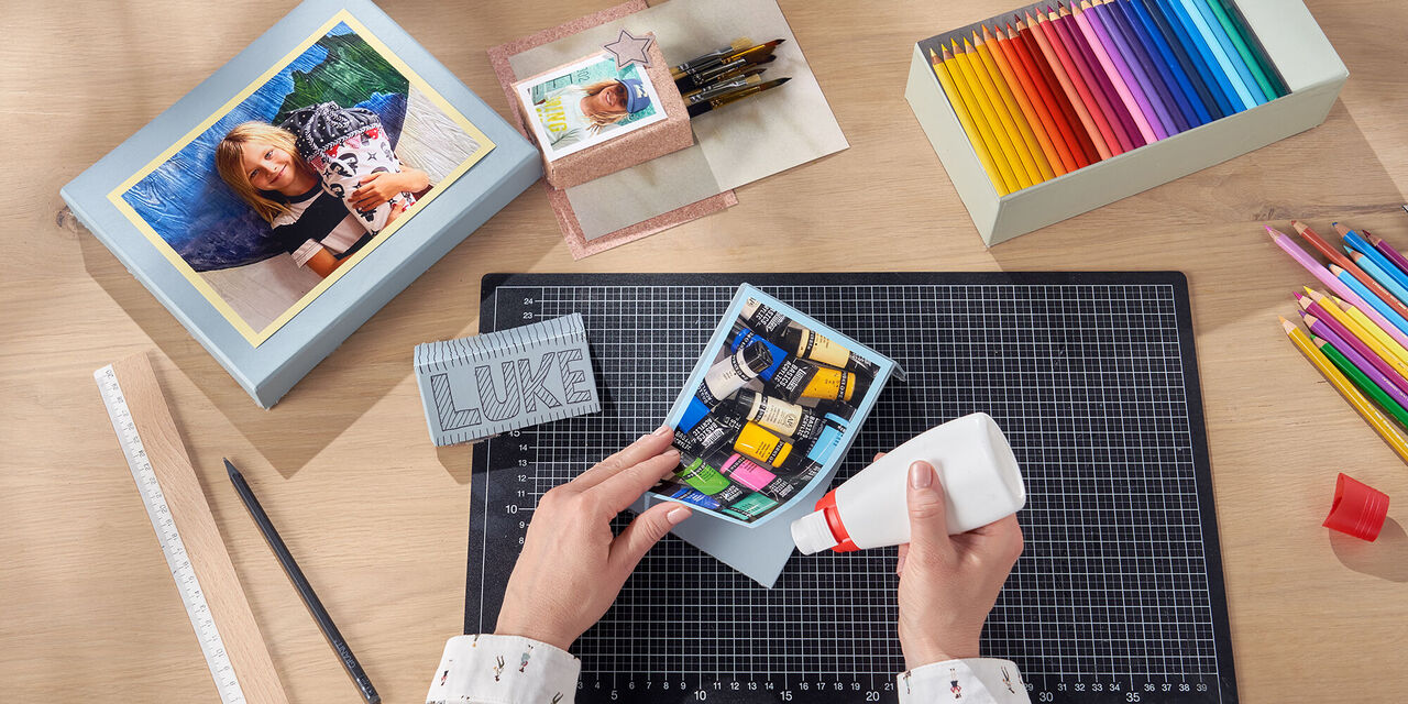 Roke pritrjujejo fotografijo, na kateri so prikazane barve za ustvarjanje, na kos kartona. Na mizi so različni ročno izdelani izdelki.